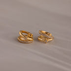The Starburst Pave Duo Huggies, gold hoops featuring a star-shaped design with small clear stones, are shown on a light beige surface.
