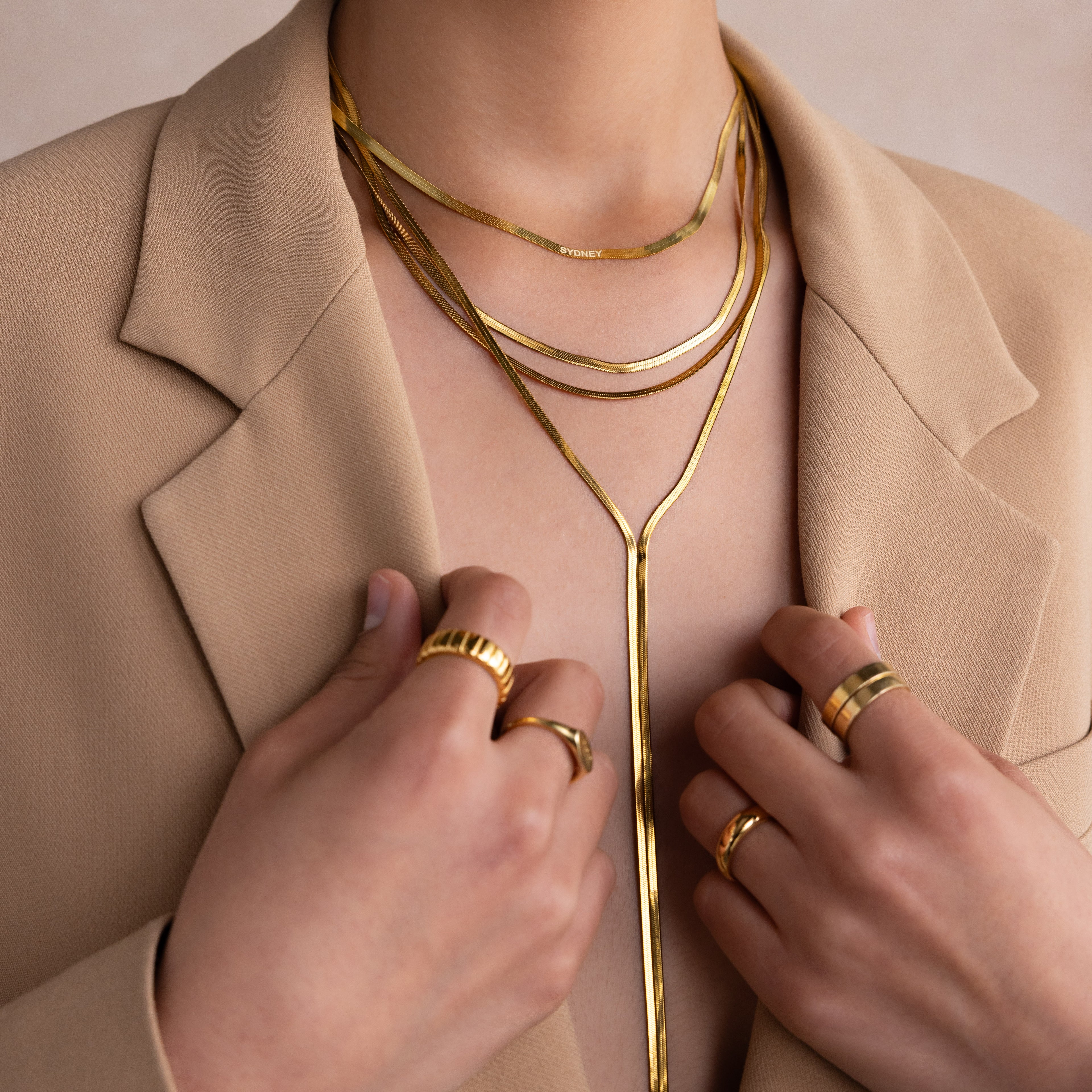 Wearing a beige blazer, the person showcases multiple gold rings and a Long Y Herringbone Chain Necklace layered with other necklaces.