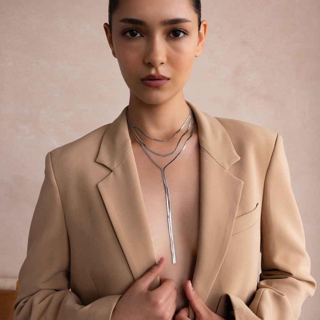 A woman in a tan blazer layers the Long Y Herringbone Chain Necklace in Silver with other silver pieces against a neutral background.