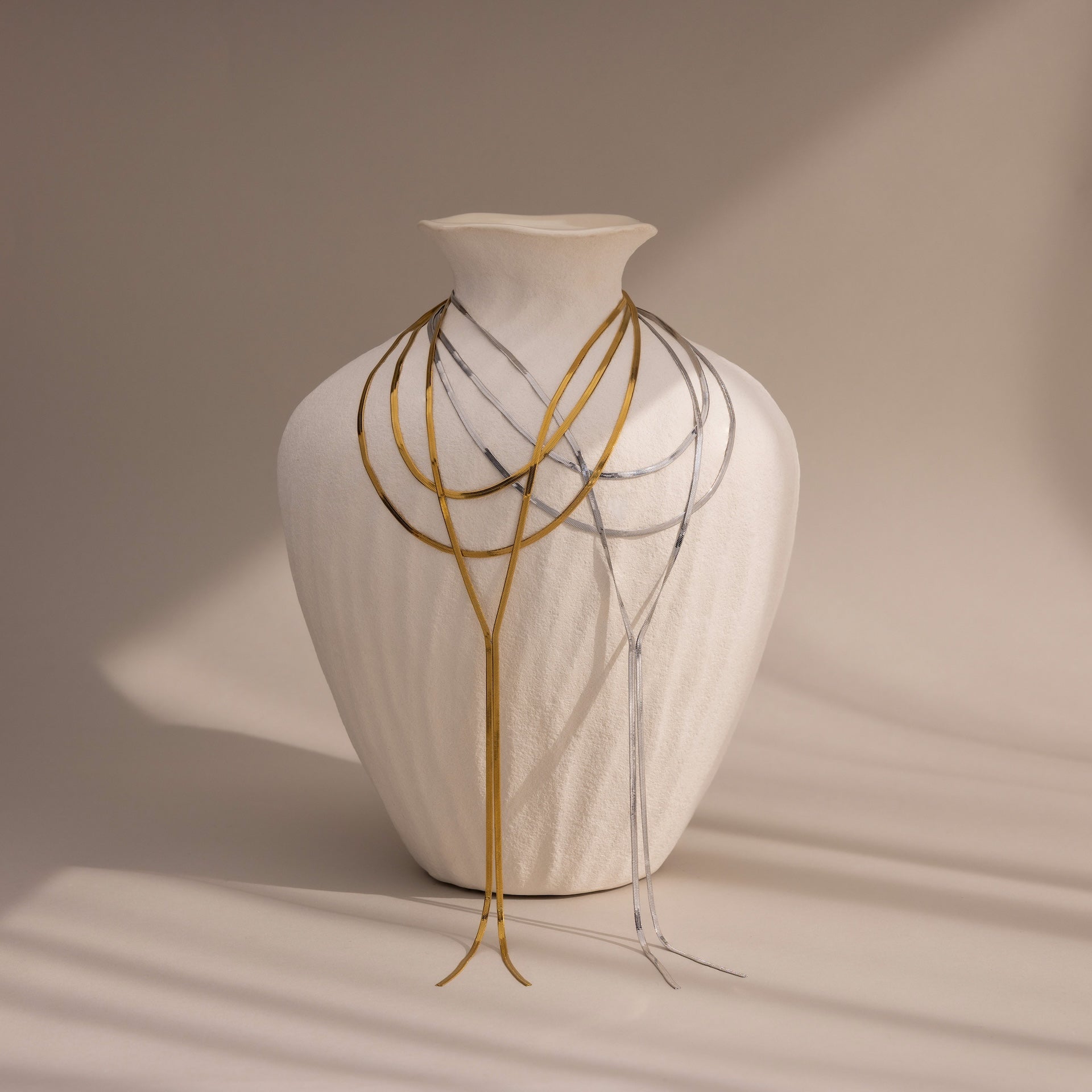 A white textured vase draped with thin gold and a Long Y Herringbone Chain Necklace in Silver, set against a neutral background.