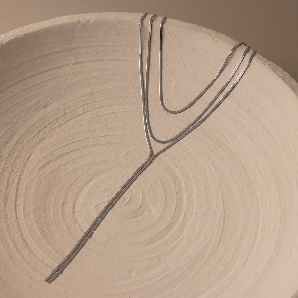 The Long Y Herringbone Chain Necklace in Silver is displayed on a textured, round beige ceramic plate with spiral patterns.