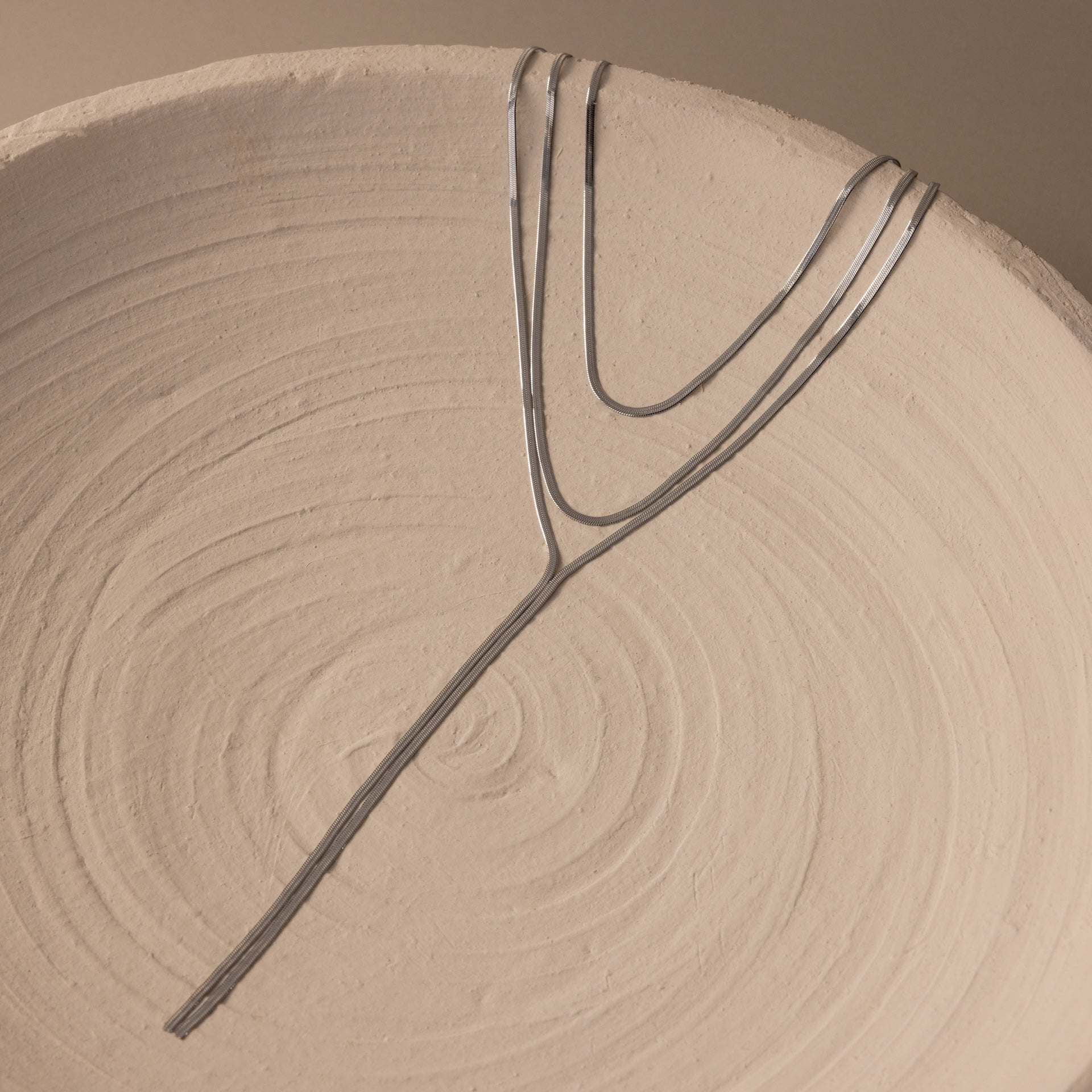 The Long Y Herringbone Chain Necklace in Silver is displayed on a textured, round beige ceramic plate with spiral patterns.