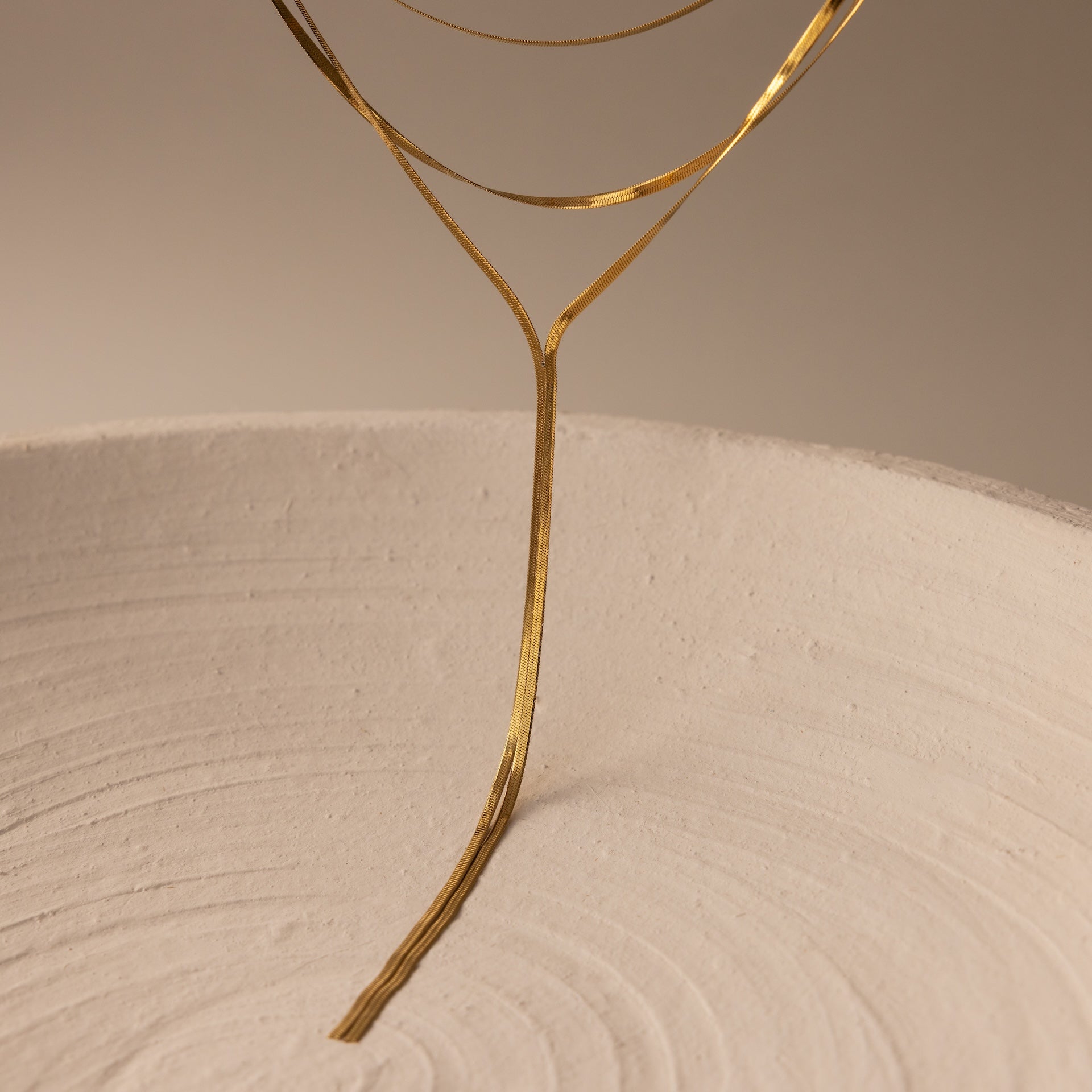 Inspired by the Long Y Herringbone Chain Necklace, this minimalist sculpture features thin gold wire extending from a textured white ceramic bowl, evoking elegant lariat-like lines in an abstract design.
