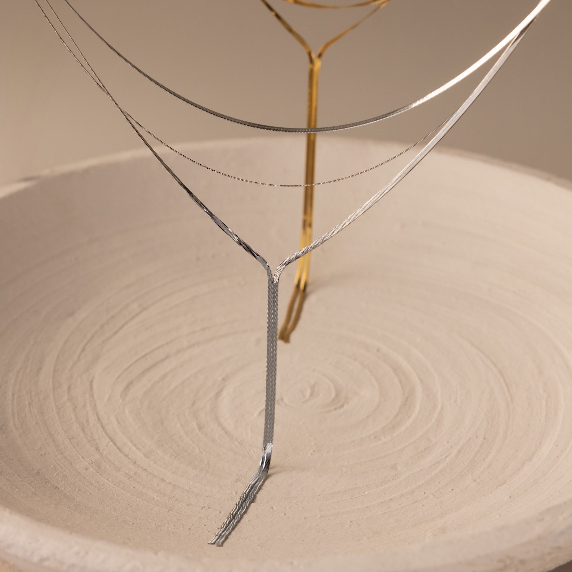Thin metal wires shaped like trees stand in a textured sand-filled dish, mirroring the refined elegance of the Long Y Herringbone Chain Necklace in Silver.