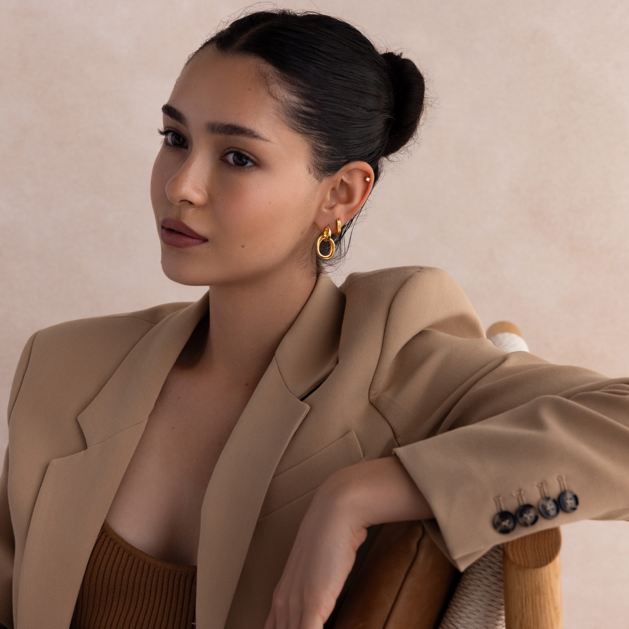 A woman with dark hair in a bun, wearing a tan blazer and gold earrings from our curated jewelry collection, sits on a chair beside our Corporate Gift Box.