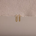 The Dainty Pave Link Earrings, featuring gold hoops with sparkling cubic zirconia stones, hang on a textured, torn beige paper set against a neutral background.