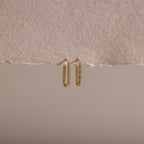 The Dainty Pave Link Earrings, featuring gold hoops with sparkling cubic zirconia stones, hang on a textured, torn beige paper set against a neutral background.