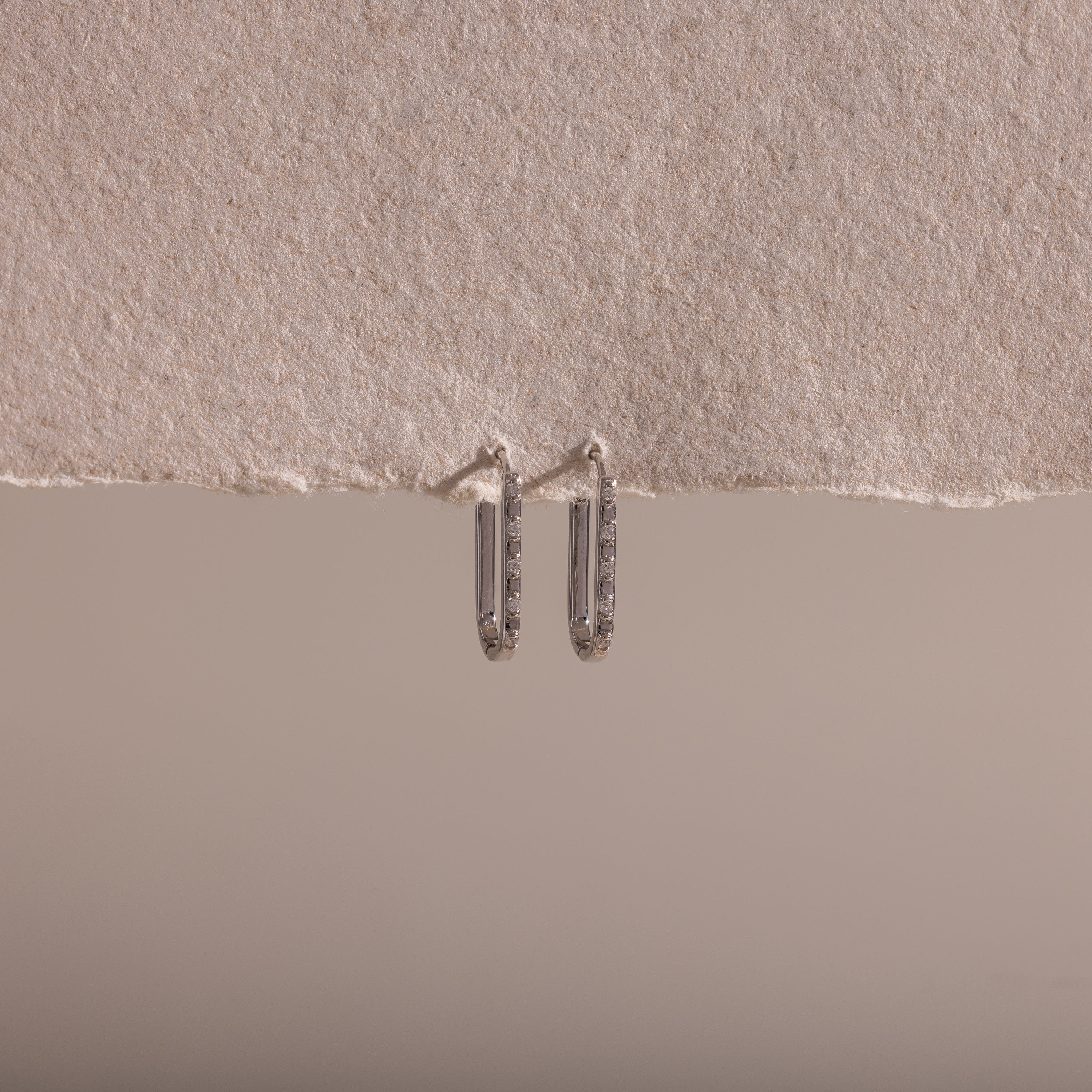 The Dainty Pave Link Earrings, featuring a minimalist silver hoop design, hang from a textured, torn edge of beige paper set against a neutral background.