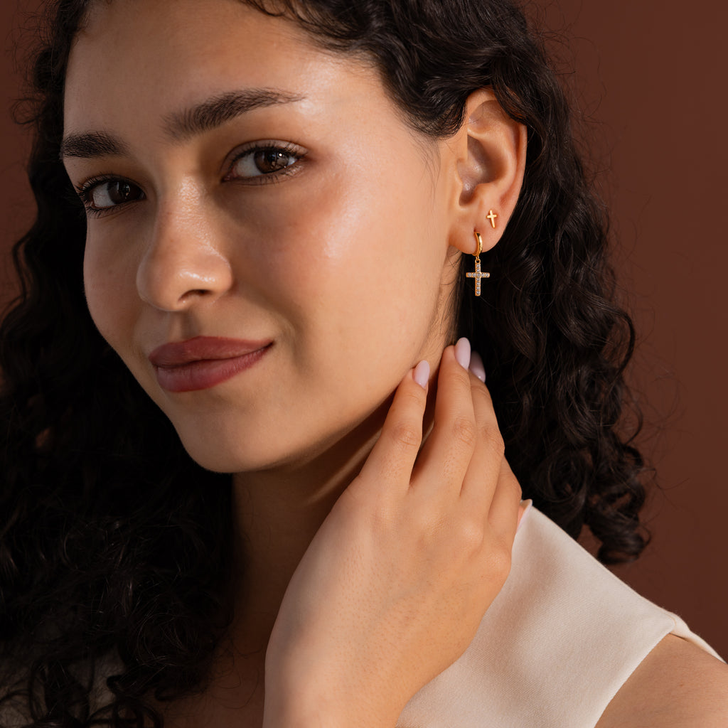 Model wearing gold huggie hoop earrings with dangling pave cross charms, stacked with dainty cross stud earrings.