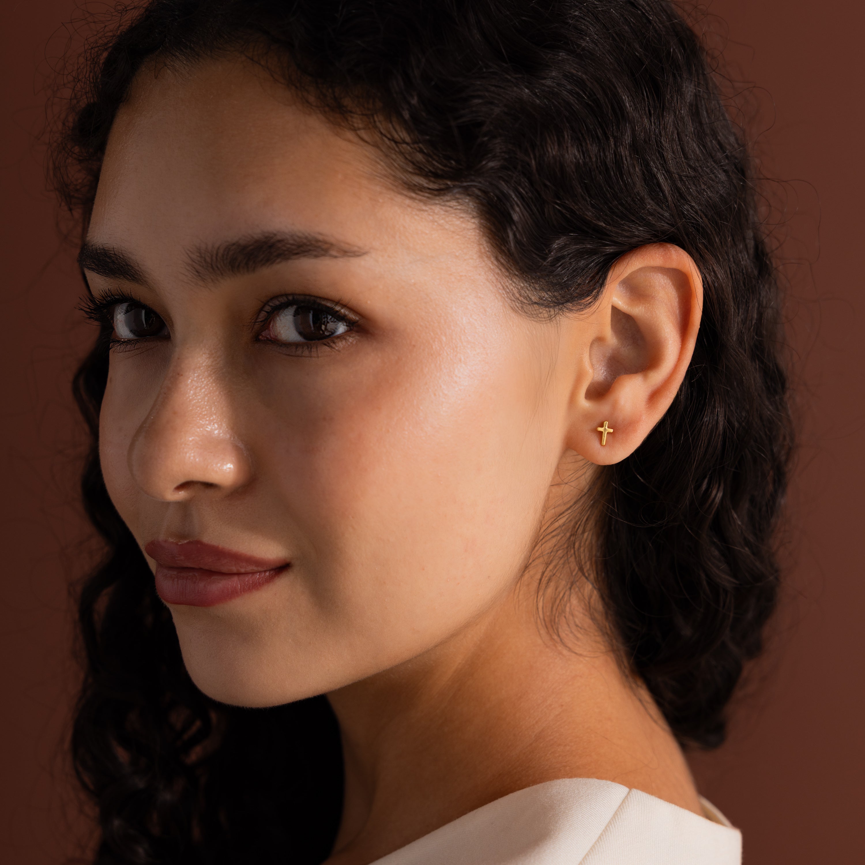 Model wearing a small gold cross stud earring, shown in a close-up portrait against a warm brown background.