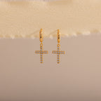Gold huggie hoop earrings with dangling pave cross charms, hanging on a neutral fabric trim.