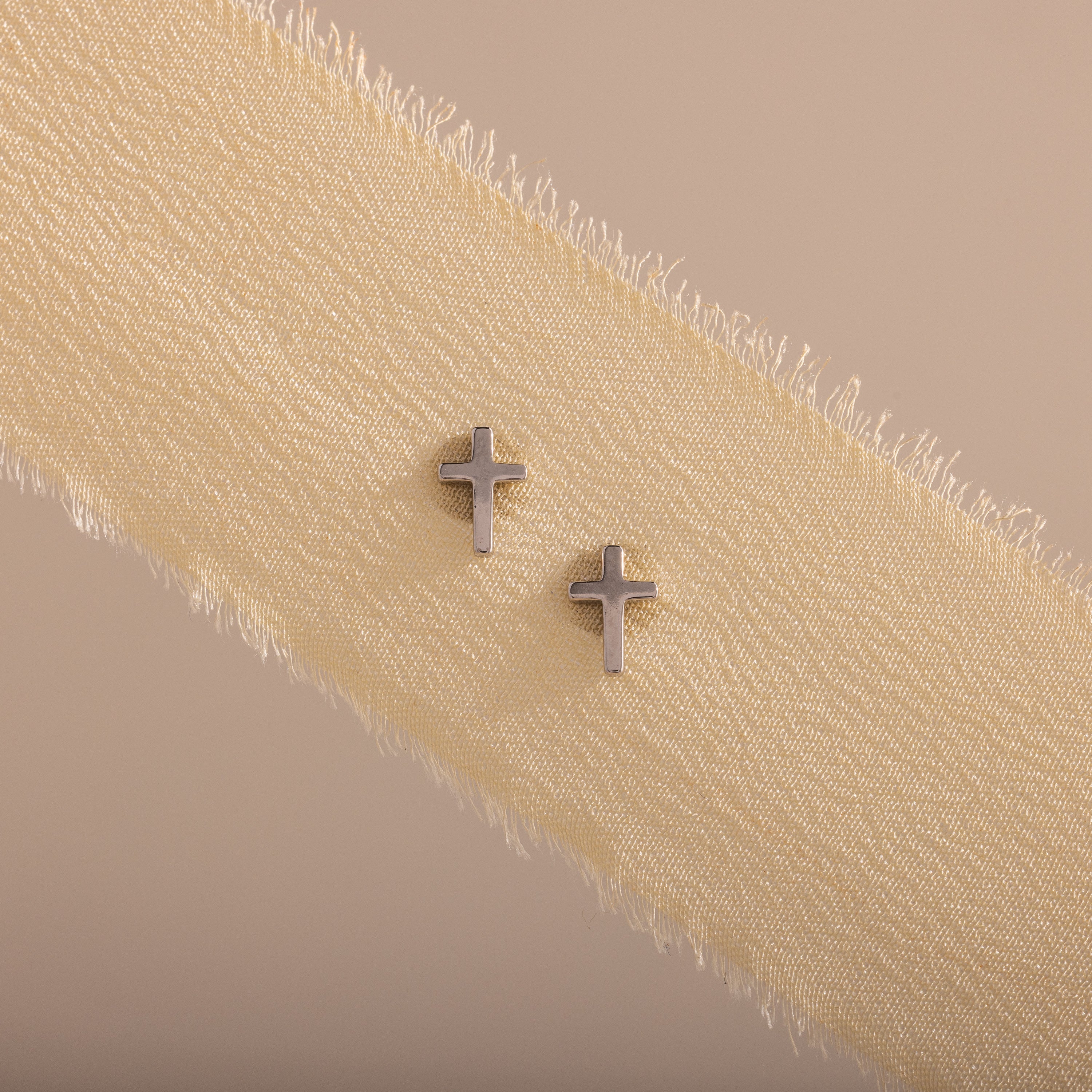 Dainty silver cross stud earrings displayed on a neutral ribbon fabric.