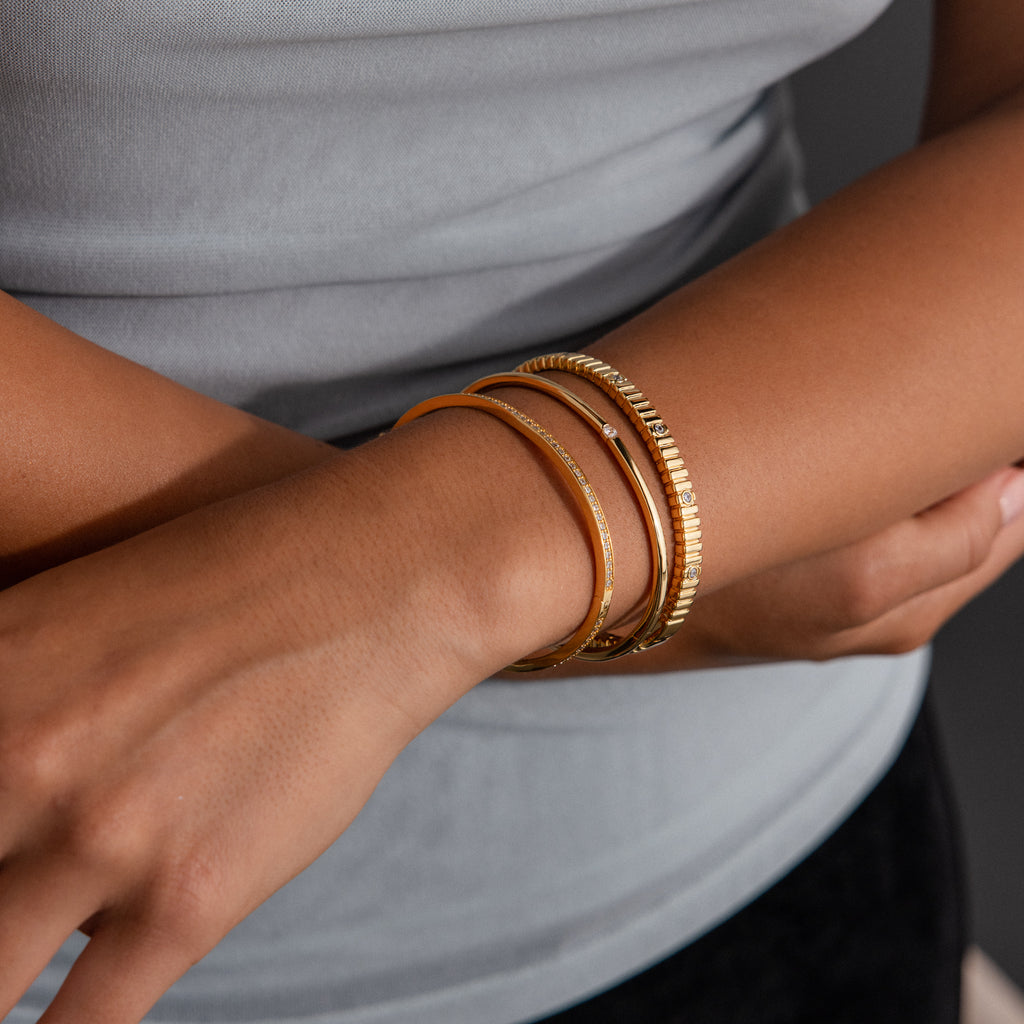 A person wears three gold bangles on their wrist, including the Ribbed Diamond Bangle, with hands gently crossed.