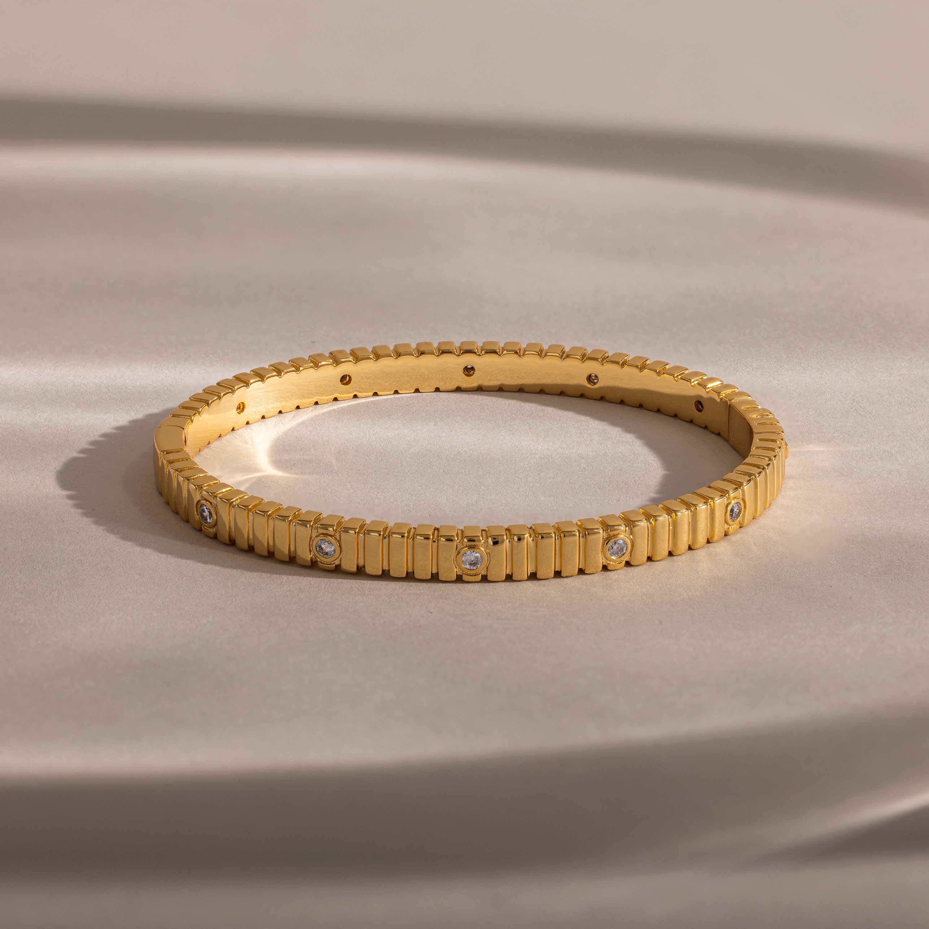 The Ribbed Diamond Bangle in 18K Gold features grooved edges and sparkling diamonds, elegantly displayed on a soft beige surface.