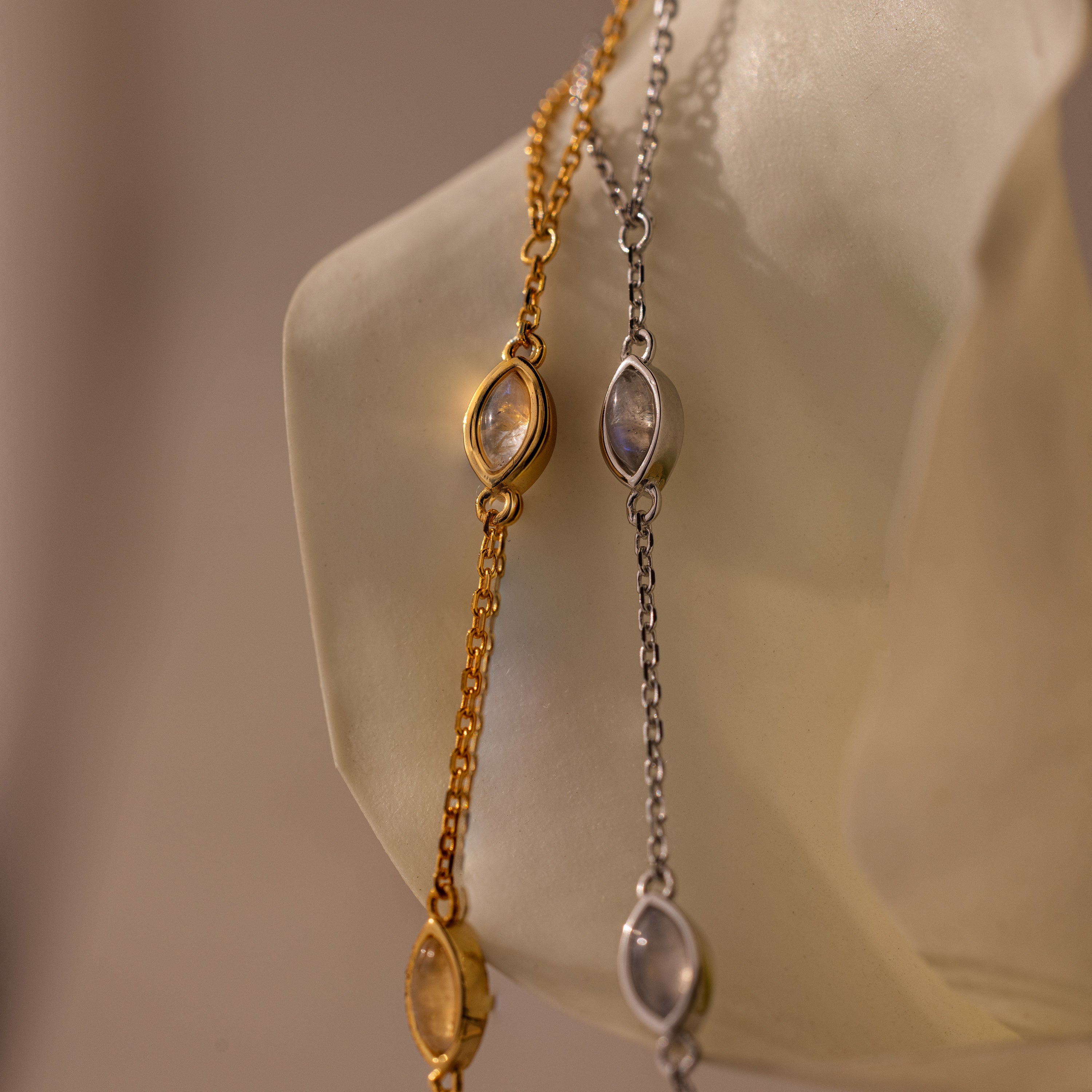 The Moonstone Hand Chain Bracelet, featuring gold and silver chains with oval moonstones, is elegantly displayed on a translucent hand-shaped stand for a celestial jewelry touch.