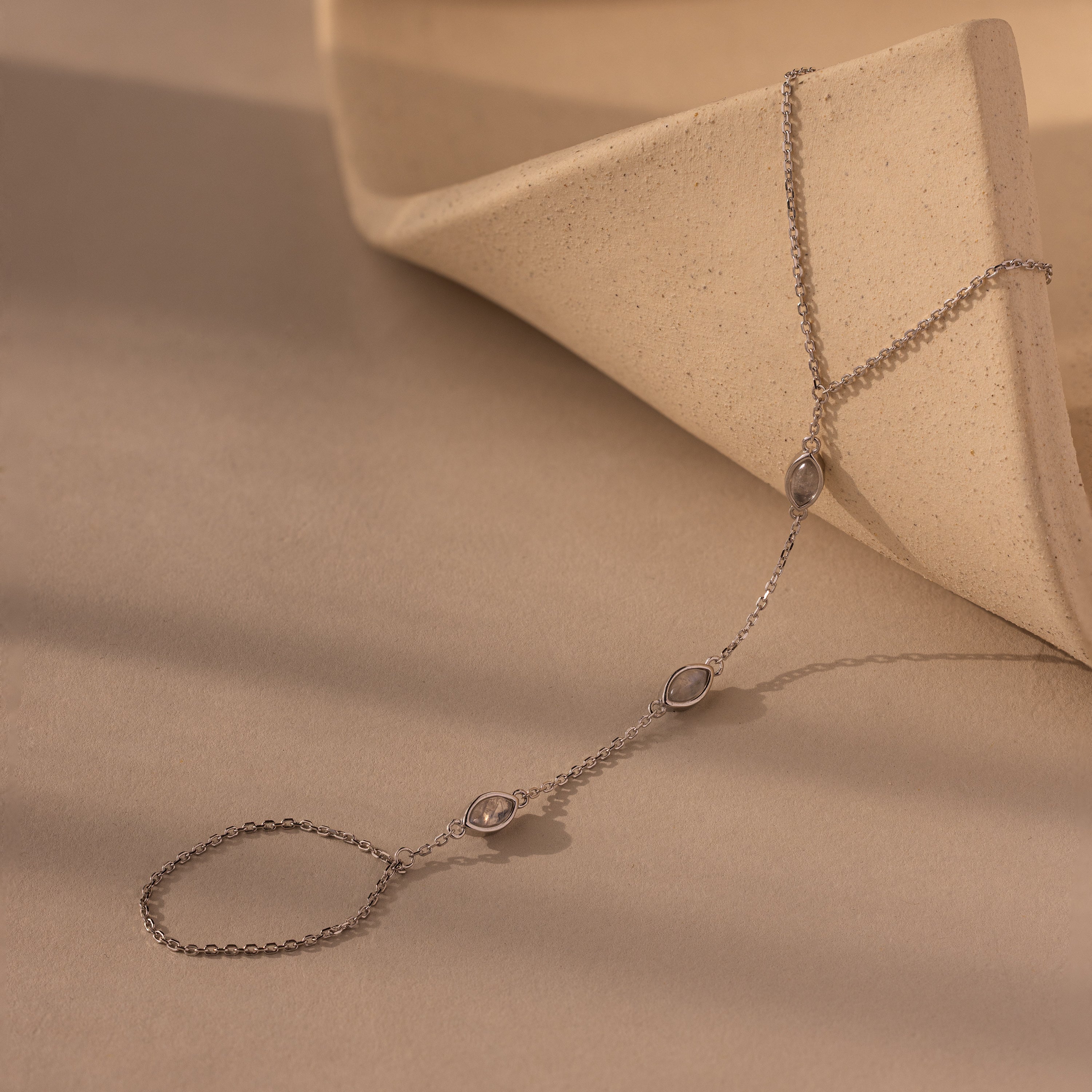 The Moonstone Hand Chain Bracelet features three oval moonstones on a delicate silver chain, displayed on a curved beige surface—a celestial accessory that brings subtle elegance to any outfit.