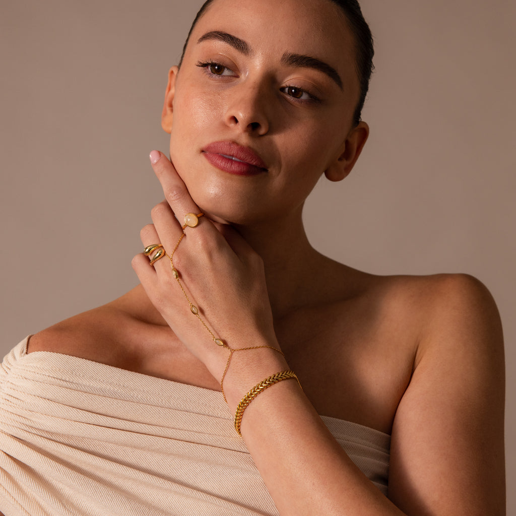 A woman with slicked-back hair wears the Moonstone Hand Chain Bracelet with celestial charm and an off-shoulder beige top, gazing thoughtfully to the side.