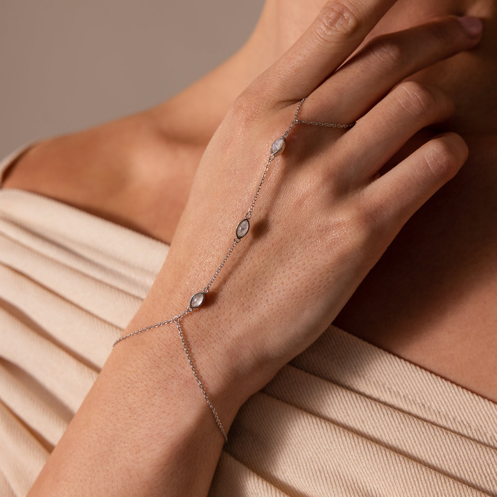 The Moonstone Hand Chain Bracelet features three delicate gemstones draping gracefully over a woman's hand and finger, beautifully displayed against a beige fabric background.