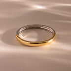 The Mixed Metal Bangle is a minimalist jewelry piece featuring silver and gold halves resting on a light, softly textured surface.