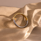 The Mixed Metal Bangle, a minimalist jewelry piece in gold and silver tones, is elegantly displayed on a textured beige wavy surface.