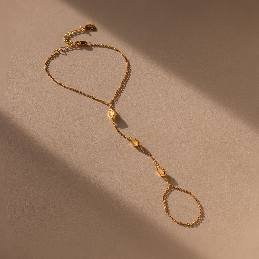 The Moonstone Hand Chain Bracelet features three delicate oval accents and is shown in soft light on a beige surface.