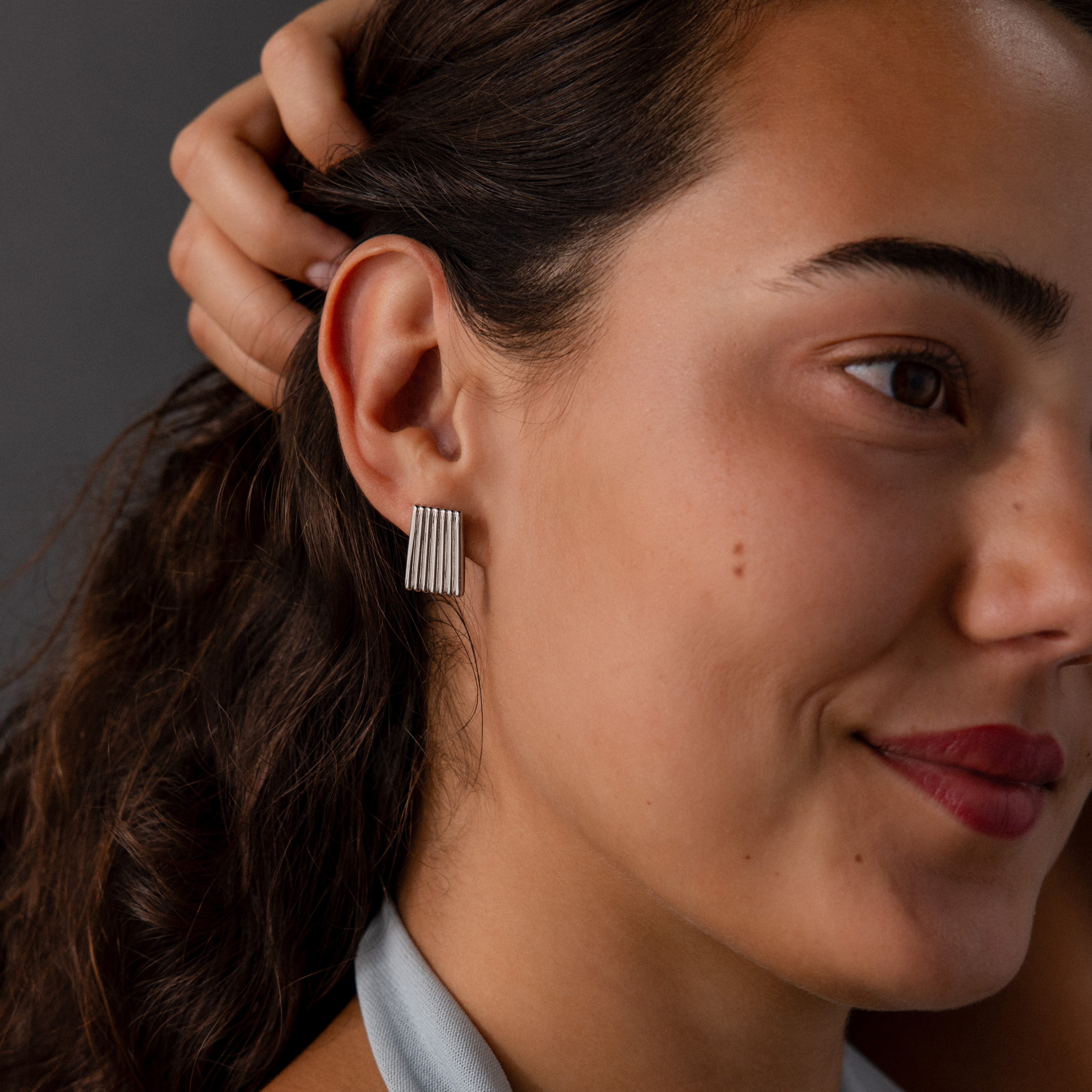 A woman smiles gently with her hand near her ear, wearing Ribbed Earrings in White Gold featuring a rectangular, textured column design—an elegant accent of contemporary jewelry.