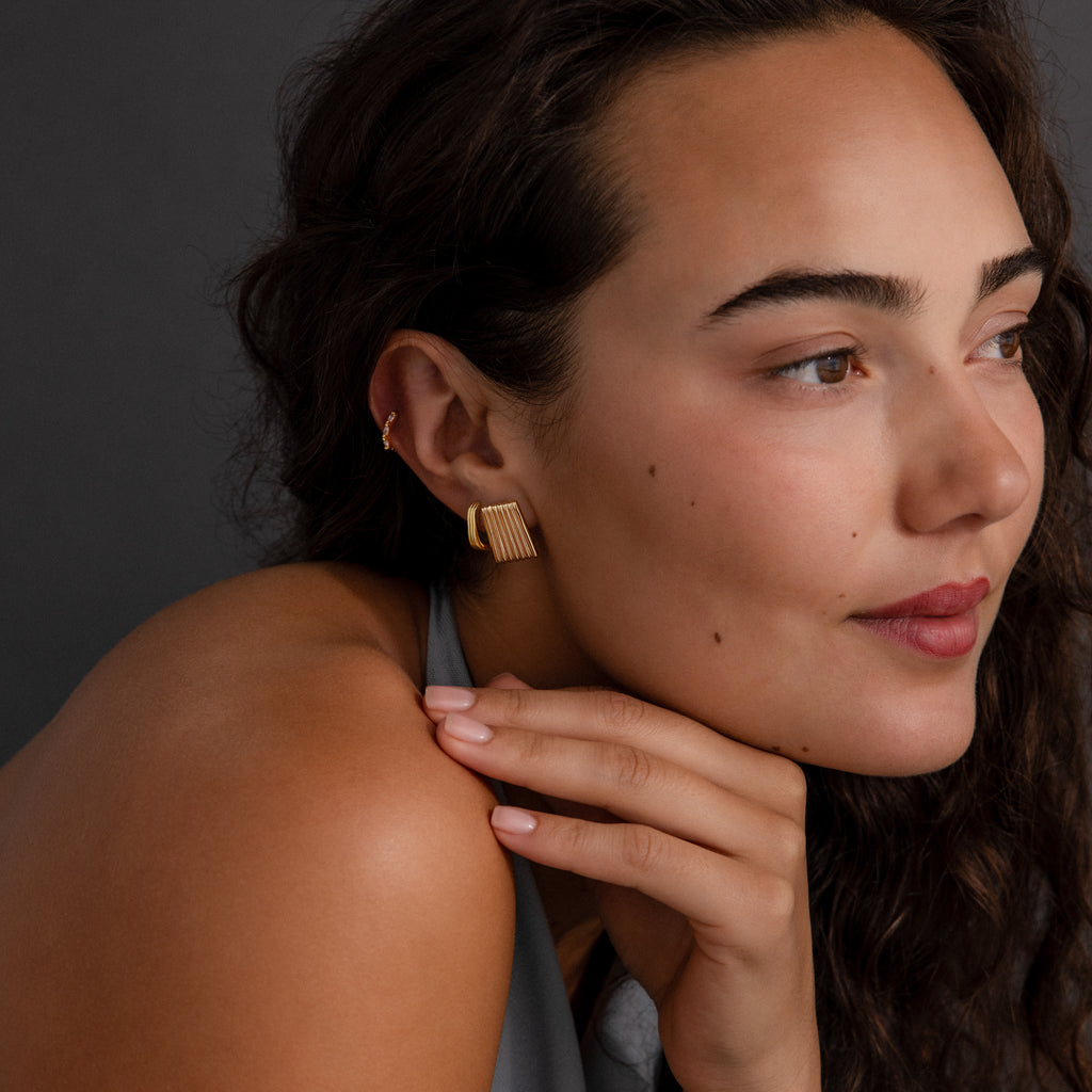 Woman with wavy brown hair wears Ribbed Earrings, resting her chin on her hand and gazing to the side.