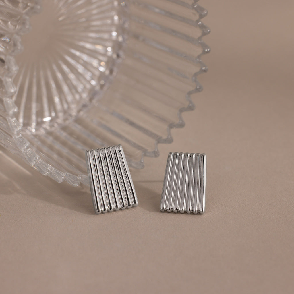 Two rectangular Ribbed Earrings in White Gold, featuring a textured column design, are displayed next to a clear ridged glass object on a beige surface.