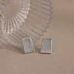 Two rectangular Ribbed Earrings in White Gold, featuring a textured column design, are displayed next to a clear ridged glass object on a beige surface.