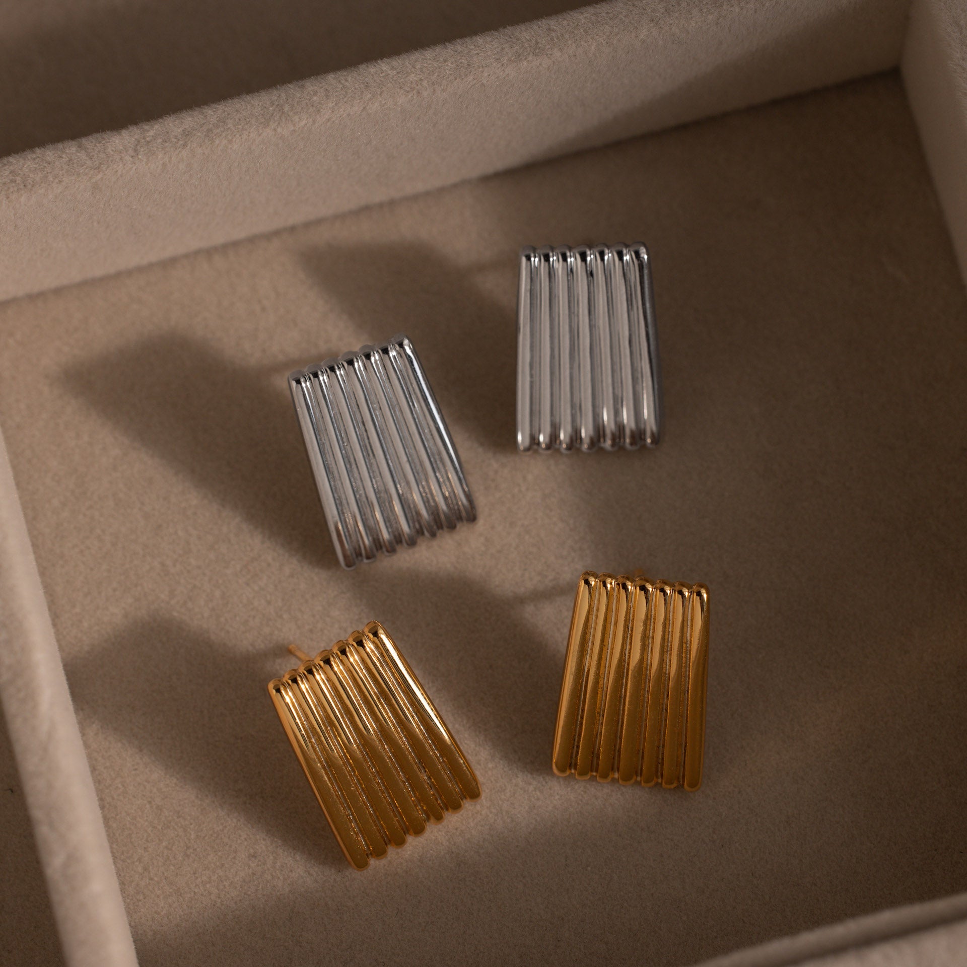 Two pairs of Ribbed Earrings—one gold, one silver—are displayed in a beige jewelry box, highlighting their sleek lines and modern style.
