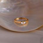 Gold heart-shaped ring with a white enamel face and small gold cross, displayed on a seashell dish.