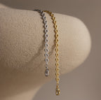 Side-by-side gold and silver Tiny Link Heart Bracelets draped over a neutral ceramic stand.