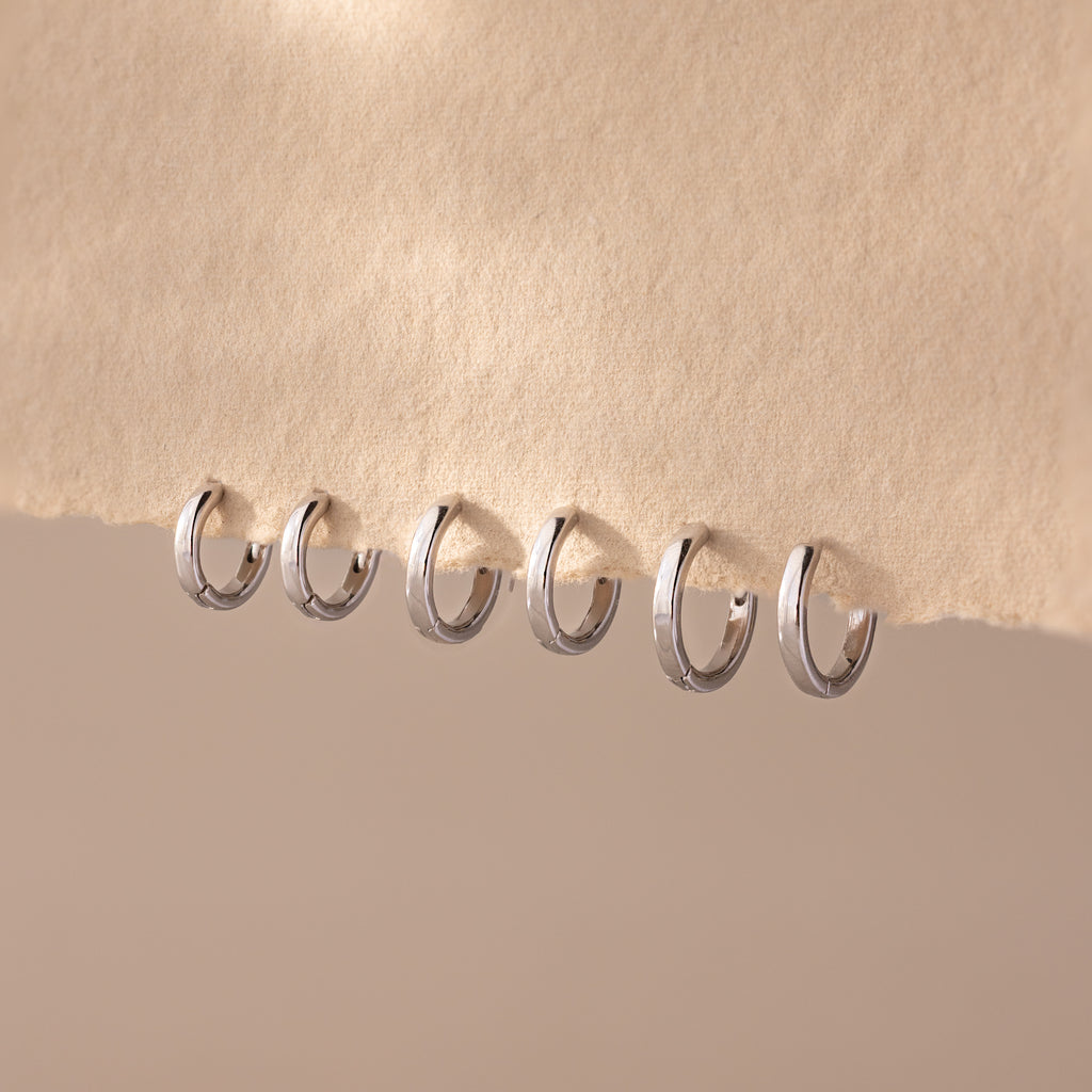 Six Dainty Huggies in silver, featuring minimalist hoop designs in increasing sizes, are lined up on a textured beige surface, highlighting their effortless style.