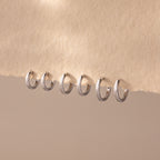Six Dainty Huggies in silver, featuring minimalist hoop designs in increasing sizes, are lined up on a textured beige surface, highlighting their effortless style.