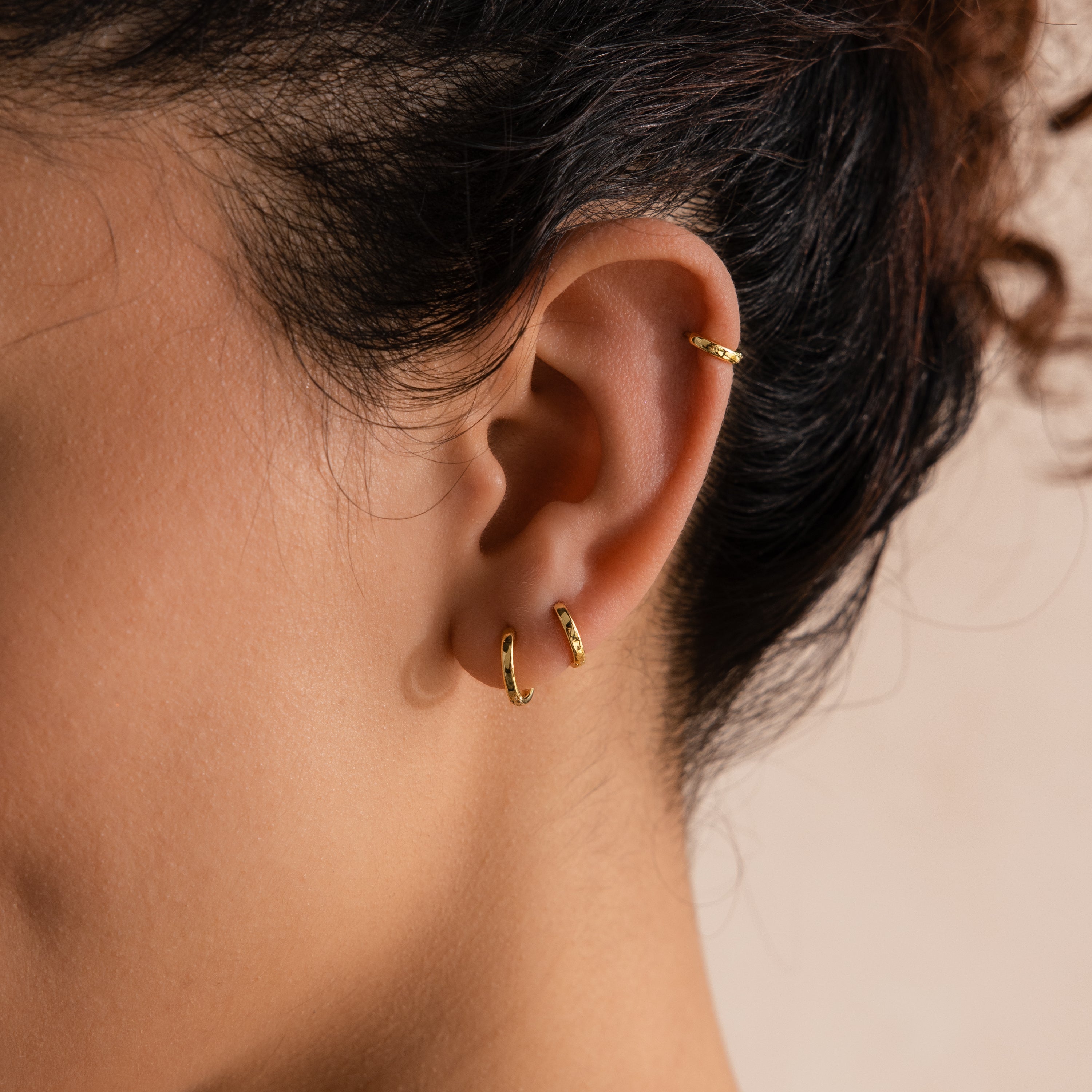 Close-up side view of model wearing three dainty gold huggie hoop earrings in her helix and lower lobe piercings.