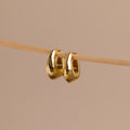 Pair of gold oval-shaped huggie earrings hanging delicately on a thin wooden rod against a neutral beige background.