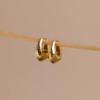 Pair of gold oval-shaped huggie earrings hanging delicately on a thin wooden rod against a neutral beige background.