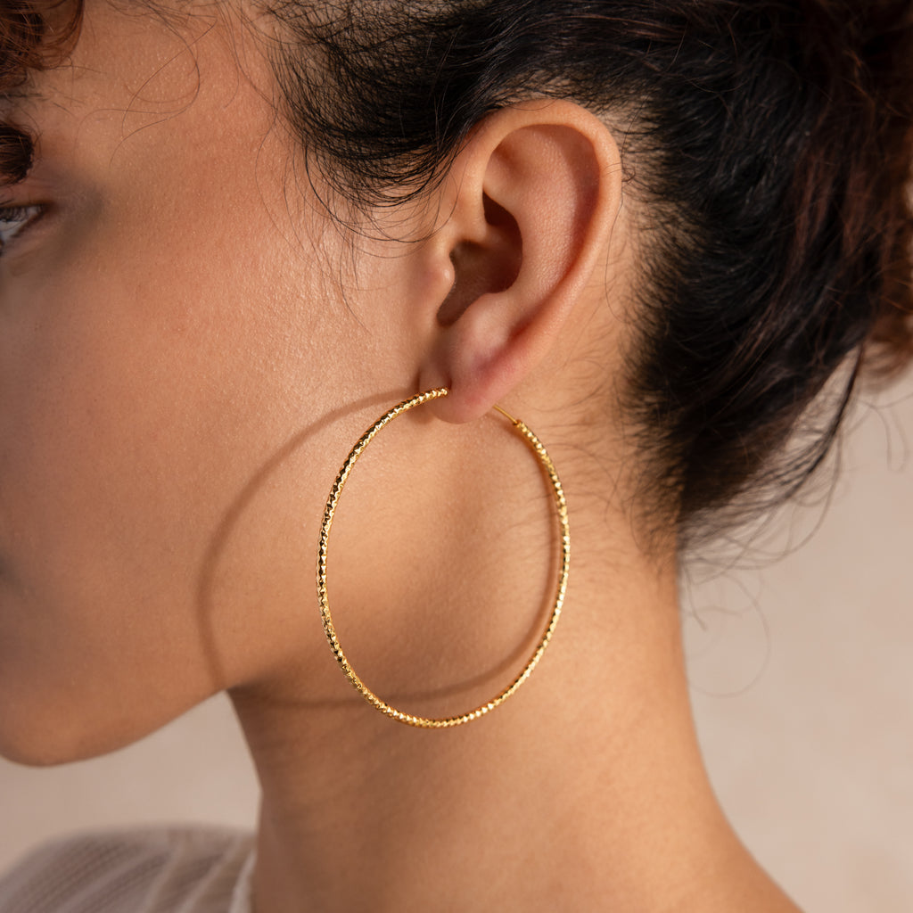 Close-up of a large textured gold hoop earring worn on the ear, highlighting its shimmering surface and lightweight design.