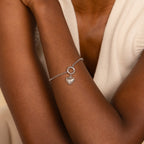 The Rolo Circle Charm Bracelet features a heart charm and a detachable hinged circle—add charms for a personalized look. Shown on the wrist with a sleeveless white top.