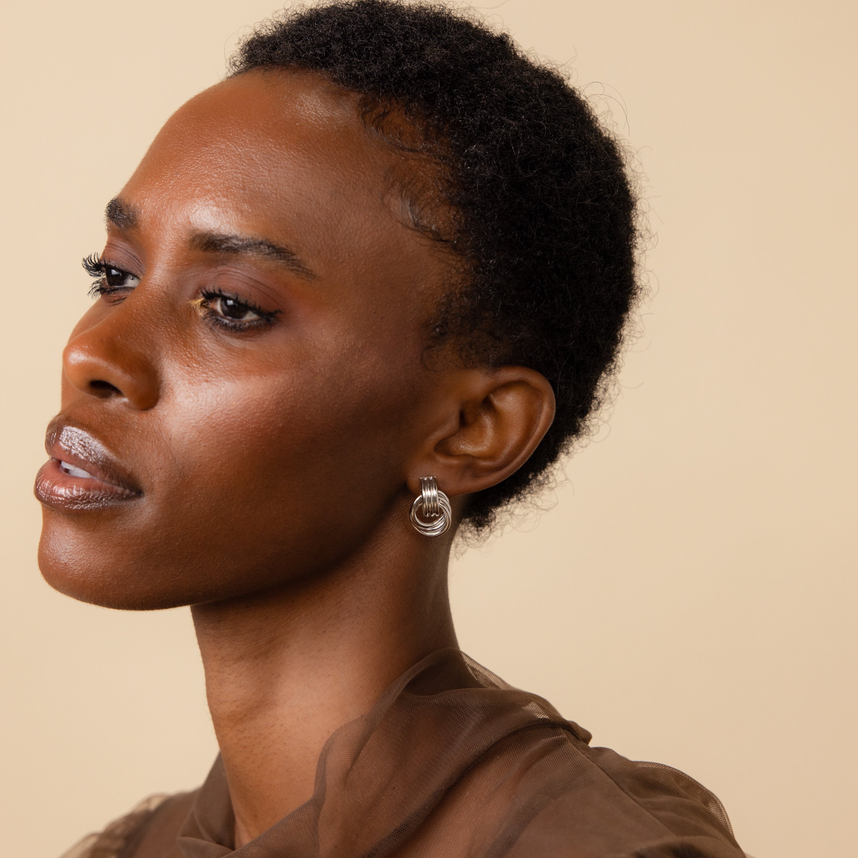 A woman with short curly hair, wearing Triple Knot Earrings and a sheer brown top, gazes left—making these earrings an inspiring anniversary gift.