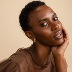 A woman poses wearing the gold-toned Mini Triple Knot Earrings, resting her hand gently on her face