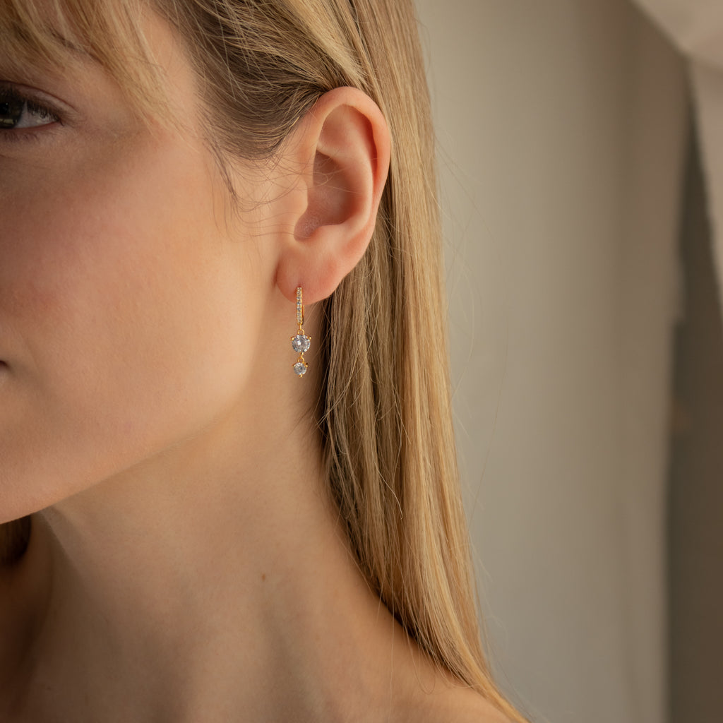 Close-up of a woman wearing Caroline Pave Drop Huggies—gold hoops with clear gemstone drops—set against a neutral background.