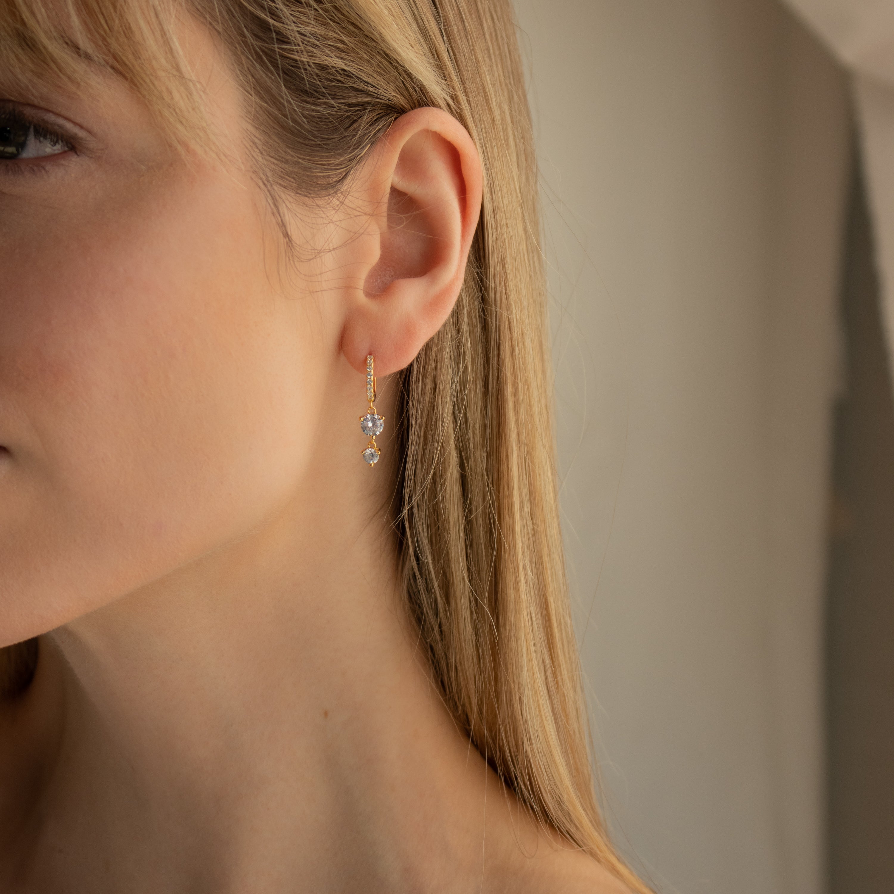 Close-up of a woman wearing Caroline Pave Drop Huggies—gold hoops with clear gemstone drops—set against a neutral background.