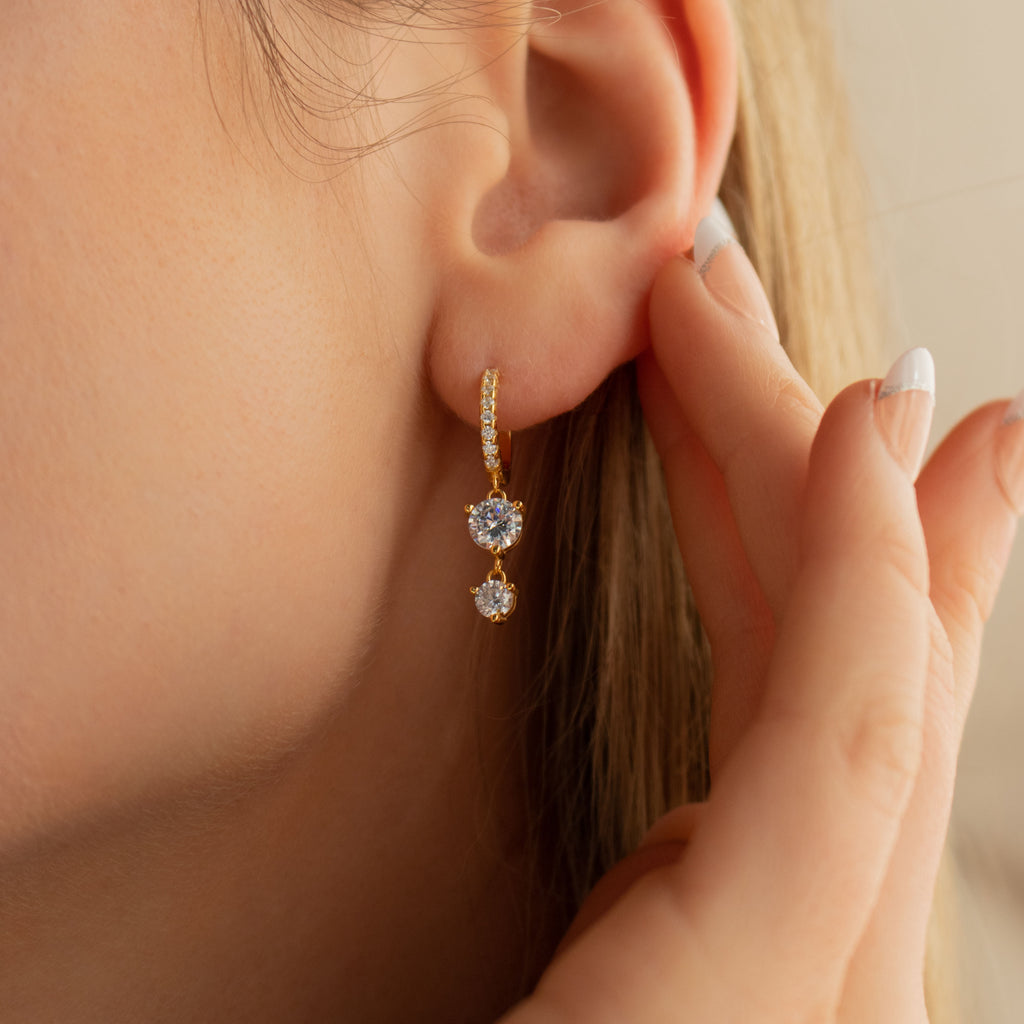 Close-up of a womans ear wearing Caroline Pave Drop Huggies, gold embellished hoops with two heart-shaped gemstones; hand touching her ear.