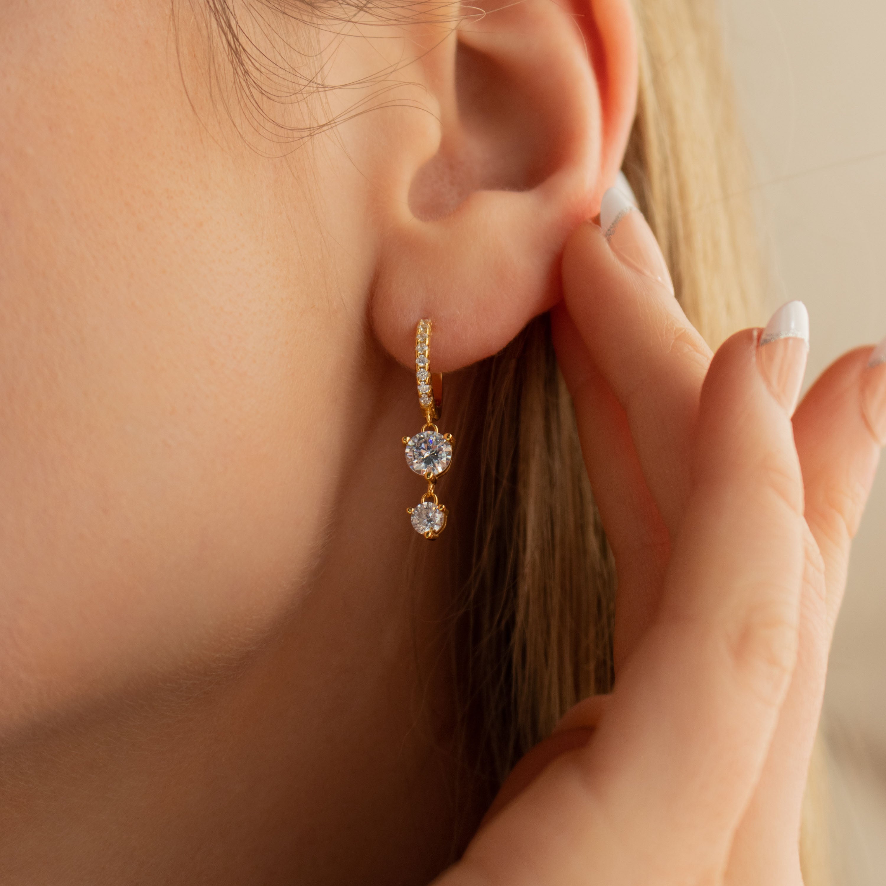 Close-up of a womans ear wearing Caroline Pave Drop Huggies, gold embellished hoops with two heart-shaped gemstones; hand touching her ear.