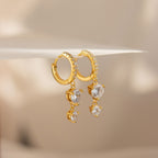 Caroline Pave Drop Huggies in gold feature dangling heart-shaped clear gemstones, shown on a soft beige background for a delicate, embellished hoop style.