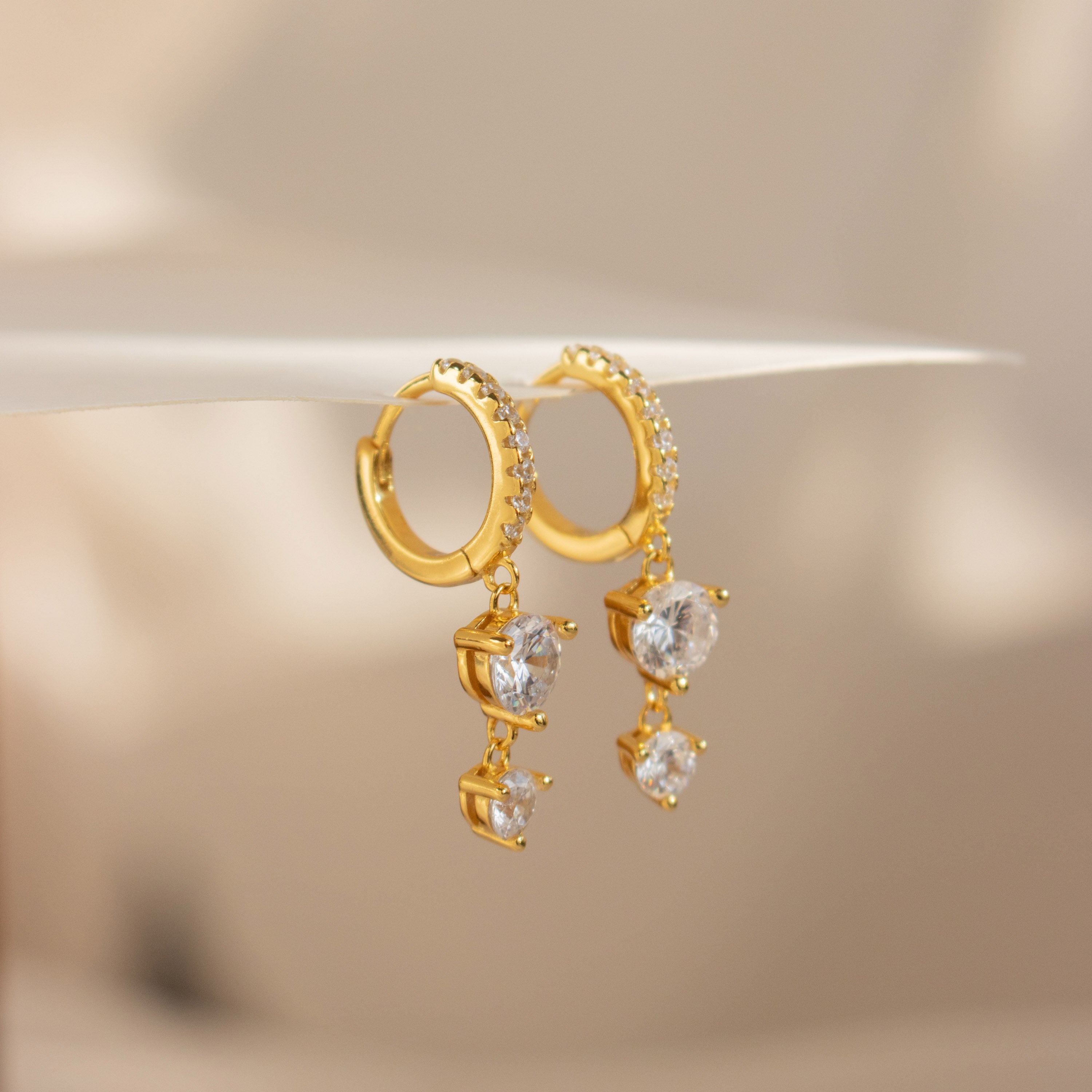 Caroline Pave Drop Huggies in gold feature dangling heart-shaped clear gemstones, shown on a soft beige background for a delicate, embellished hoop style.