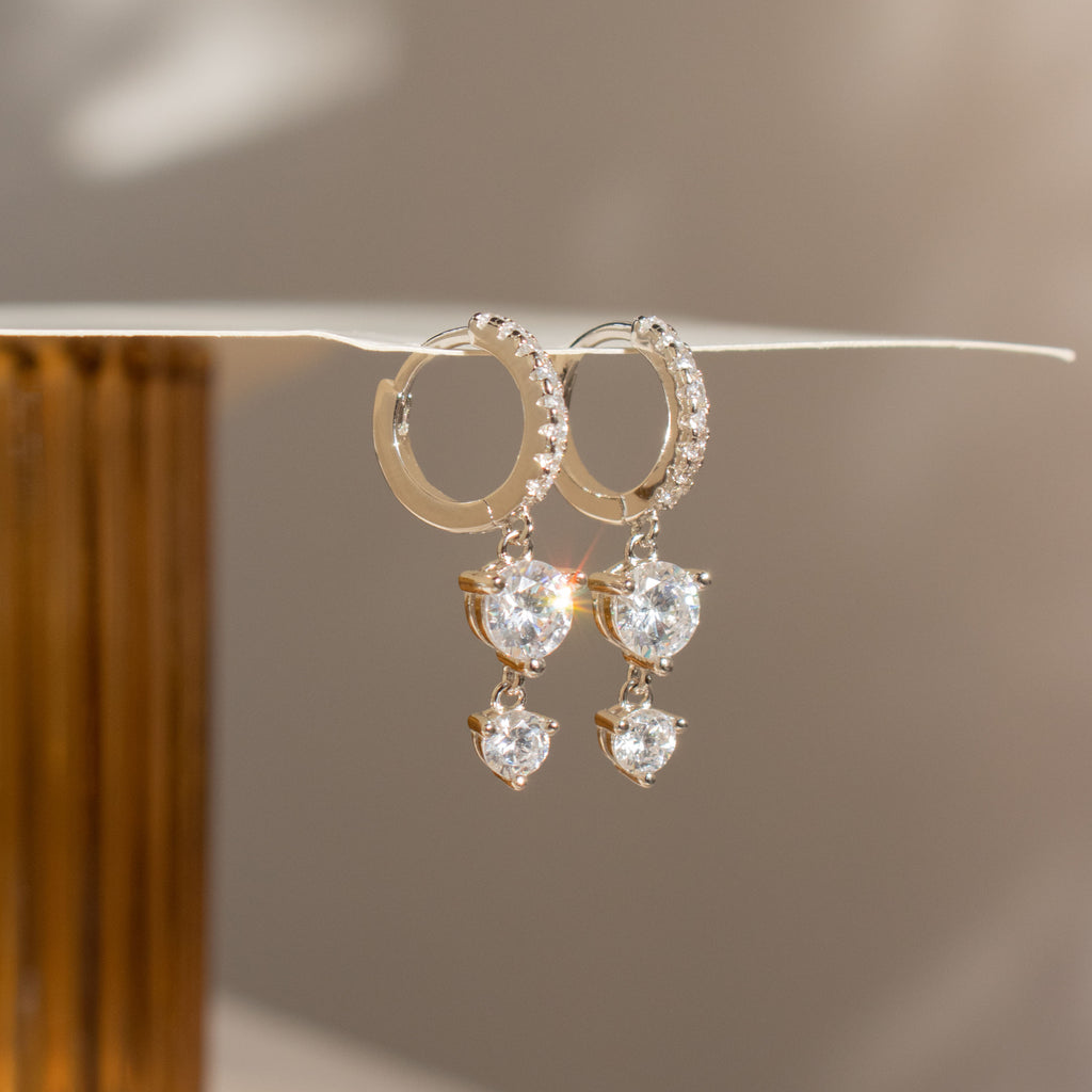 Caroline Pave Drop Huggies: sparkling gold hoops adorned with teardrop gems, showcased on a white surface.