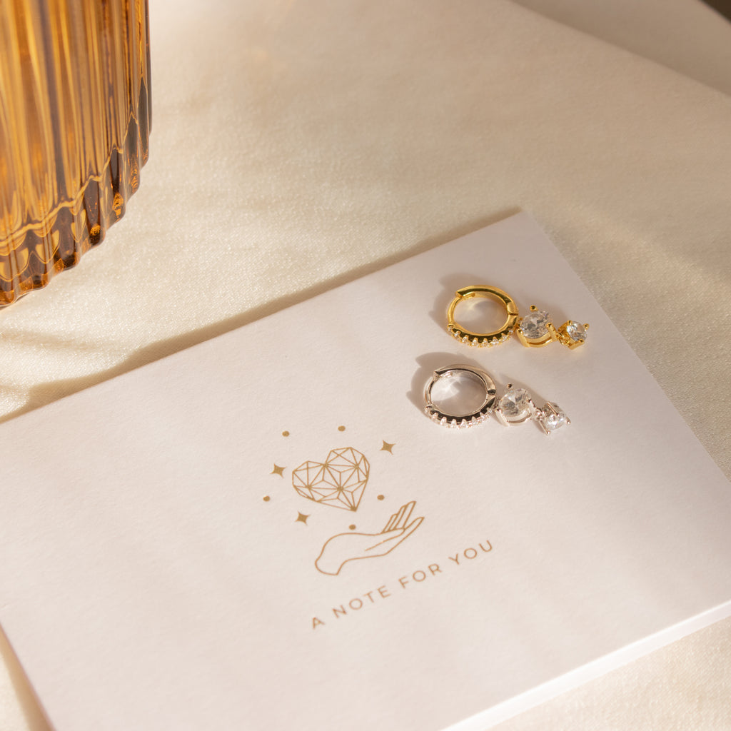 Caroline Pave Drop Huggies earrings rest on a card that says A NOTE FOR YOU, featuring a hand and heart illustration.