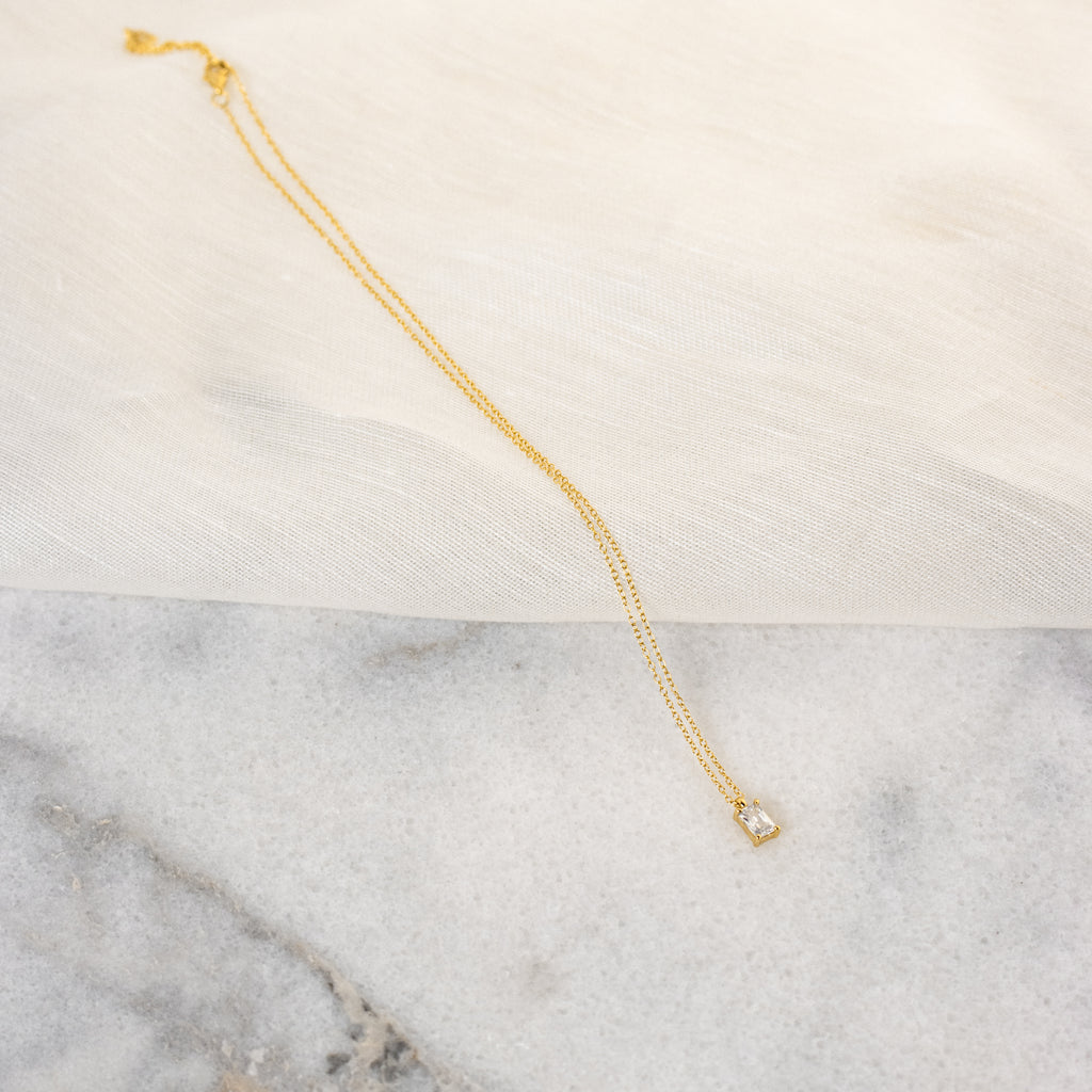 The Sage Diamond Necklace in 18K Gold features a delicate square pendant, radiating timeless elegance against a marble and fabric backdrop.